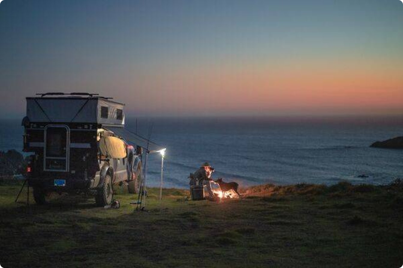 Overlander camper parked on coastal bluff at dusk with person and dog by campfire, ocean backdrop.