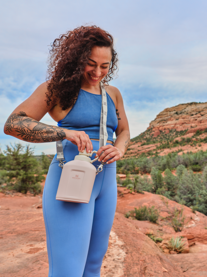 Woman smiling outdoors in Sedona, opening a Stanley 1913 cream jug with crossbody strap, wearing blue activewear.