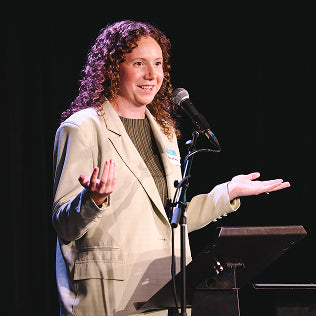 Woman speaking at a podium with a microphone on a dark stage, wearing a light blazer.