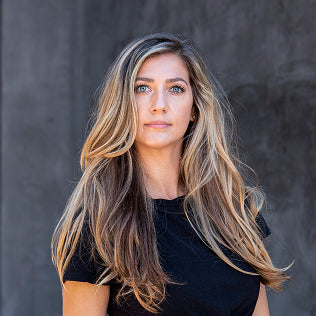 Woman with long blonde highlighted hair and blue eyes in a black top against a concrete wall.