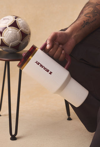 Stanley 1913 white tumbler with maroon handle and gold lid, held by a person seated near a soccer ball.