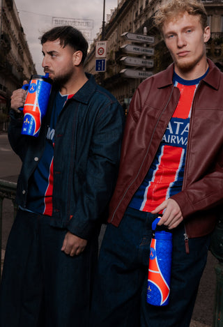 Two men in PSG jerseys on a Paris street, one drinking from a Stanley 1913 blue PSG tumbler, other holding matching bottle.