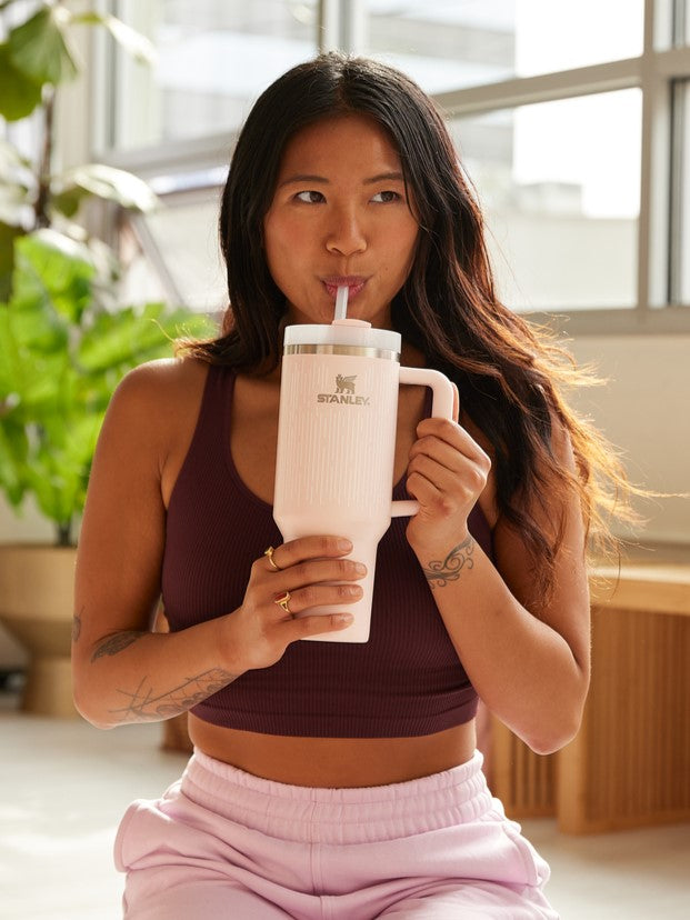 Woman drinking from a light pink Stanley 1913 tumbler with handle and straw, wearing a burgundy top indoors.